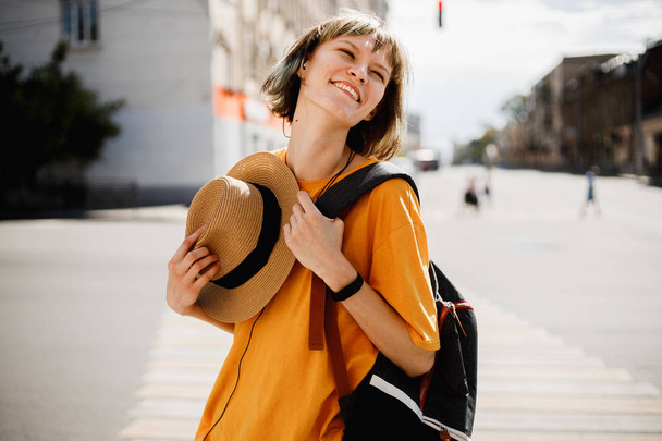 Funny young girl with headphones dressed in a yellow t-shirt holds a straw hat in her hand standing in a city street on a summer sunny day - 写真・画像