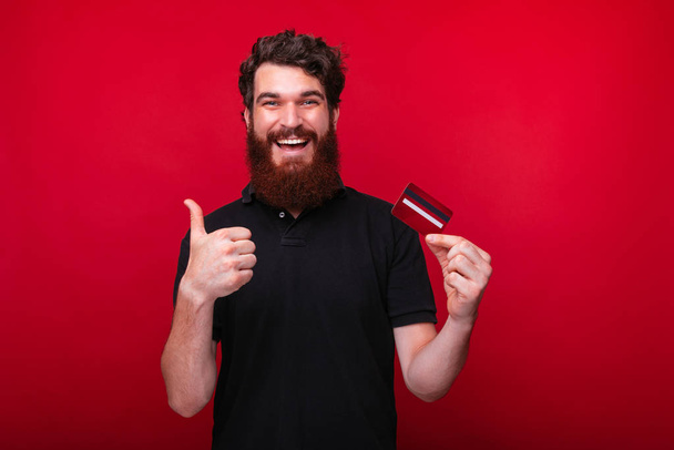 Portrait of bearded man holding a credit card, while showing thu - Foto, Bild