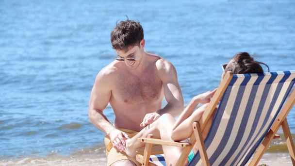 vrolijke jonge man die zonnebrandcrème aanbrengt op het been van jonge vriendin zonnebaden in Chaise longue - Video