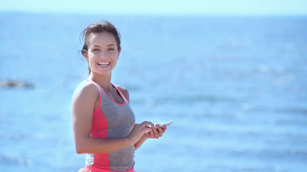 fröhliche junge Frau in Sportbekleidung mit Smartphone, blickt in die Kamera und zeigt Siegergeste - Filmmaterial, Video