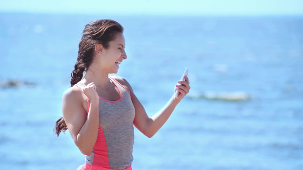 привлекательная молодая женщина, использующая смартфон и улыбающаяся, показывая жест победителя
  - Кадры, видео