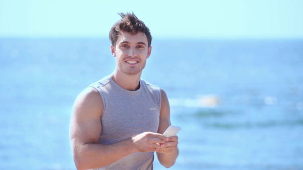 ευτυχισμένος νέος άνθρωπος σε αθλητικά είδη χρησιμοποιώντας smartphone στην παραλία και δείχνοντας Ναι χειρονομία - Πλάνα, βίντεο