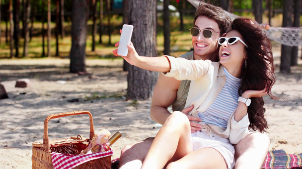 χαρούμενο νεαρό ζευγάρι παίρνοντας selfie στο smartphone και δείχνοντας χειρονομίες νίκης ενώ αναπαύεται στην παραλία - Πλάνα, βίντεο