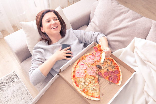 Ritratto di giovane donna che ride sdraiata sul divano, mangia pizza e tiene il tono arancione del telefono
 - Foto, immagini