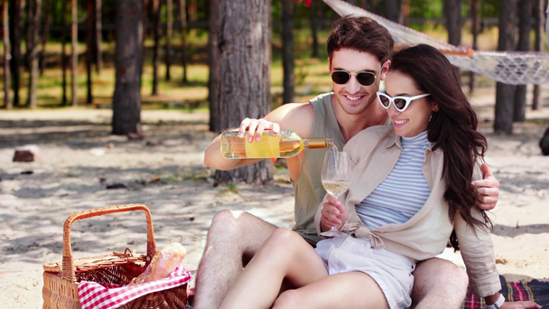 przystojny młody człowiek przytulanie i Całowanie dziewczynę i nalewanie białego wina w szklance podczas odpoczynku na plaży - Materiał filmowy, wideo