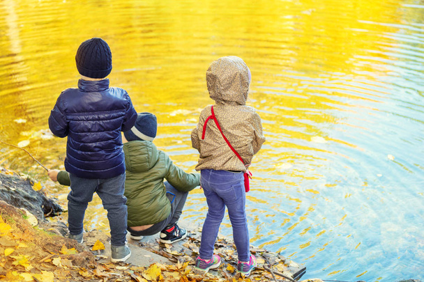 Three children friends playing fishing with sticks near pond in beautiful golden fall park. Little siblings having fun near lake or river in autumn. Happy childhood and kid outdoor recreation concept - Fotografie, Obrázek
