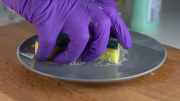A hand in blue protective gloves squeezes a yellow-green sponge and washes dishes. The foam bubbles and drains onto a gray plate. Slow motion. Housekeeping with quality detergent - Footage, Video