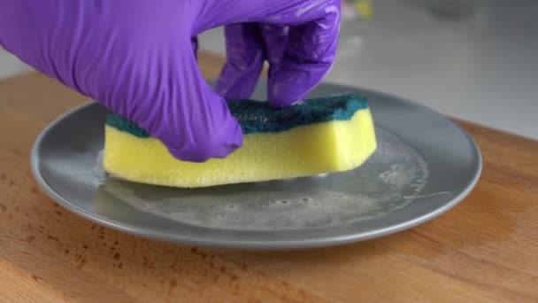 Een hand in blauwe handschoenen wast een grijze plaat met een geel-groene spons en schuimreiniger. Op een houten oppervlak. Slow Motion - Video