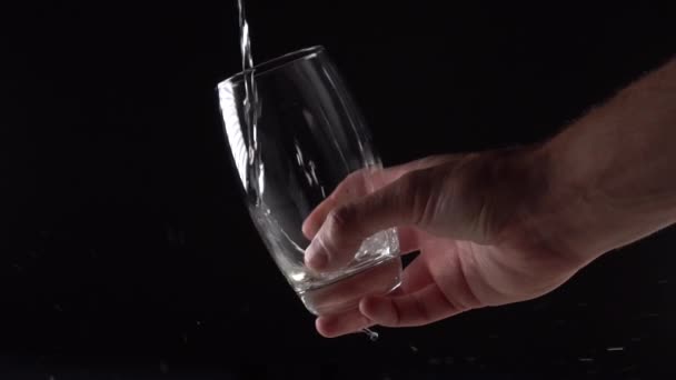 Water pours from above and fills a glass cup in a hand on a black background. Slow motion - Filmati, video