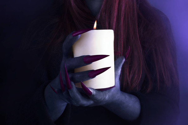 Handen van een heks met lange scherpe klauwen die een brandende kaars houden, close-up. Halloween, Black Magic concept. - Foto, afbeelding