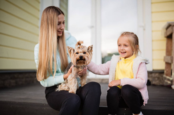 Ładna kobieta z długimi blond włosami trzymająca małego psa Yorkshire Terrier na świeżym powietrzu, następna siedząca dziewczynka. Szczęśliwa matka i córka ze zwierzątkiem na podwórku. Koncepcja rodziny - Zdjęcie, obraz