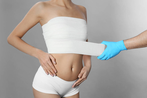 Doctor bandaging young woman after plastic surgery operation on grey background, closeup - Φωτογραφία, εικόνα