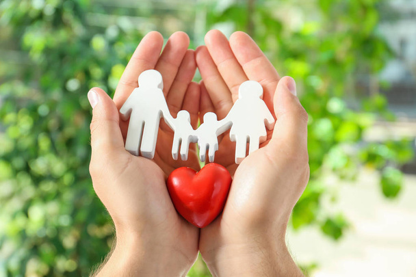 Young man holding wooden family figure and red heart in his hands against blurred background, closeup - Foto, Imagem