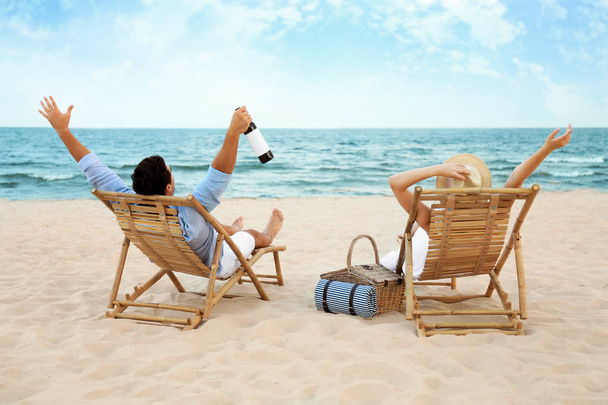 Happy young couple with wine sitting on deck chairs at sea beach - Foto, Imagem