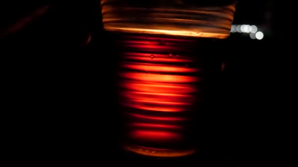 Przezroczysta plastikowa filiżanka wypełniona koniakiem jest migotanie na tle ognia w nocy - Materiał filmowy, wideo