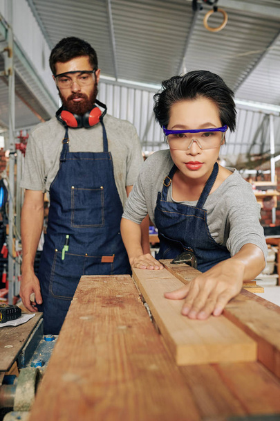 Serious professional carpenter looking at his female coworker putting wooden planks on workbench - Photo, Image