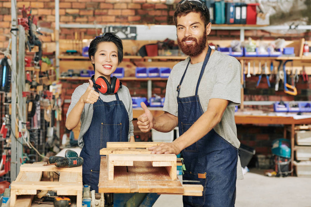 Felices carpinteros sonrientes de pie en el banco de trabajo y mostrando los pulgares hacia arriba
 - Foto, imagen