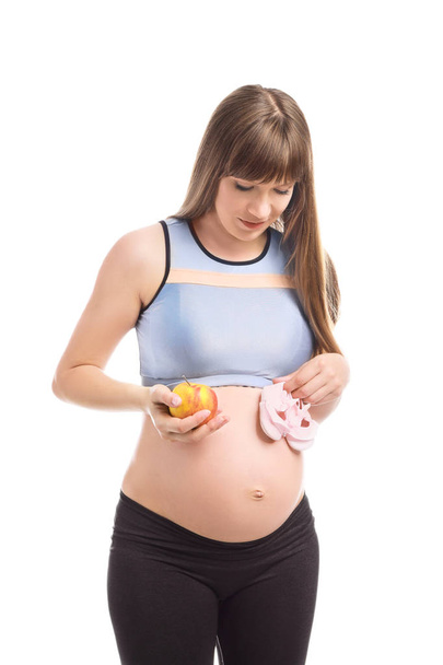 Беременная женщина с яблоком и детскими сапожками на белом фоне
 - Фото, изображение