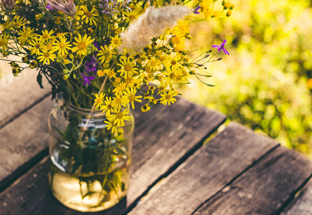 bouquet of yellow wildflowers in a glass jar on a wooden background - Фото, изображение