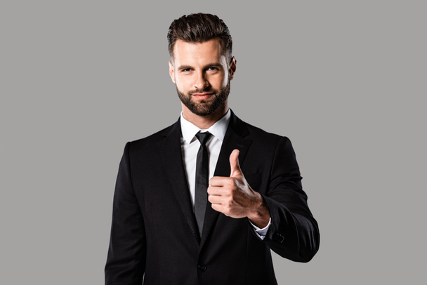 Lächelnder gutaussehender Geschäftsmann im schwarzen Anzug, der den Daumen vereinzelt auf grau zeigt - Foto, Bild