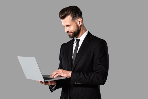 homme d'affaires beau et souriant en costume noir en utilisant un ordinateur portable isolé sur gris
 - Photo, image