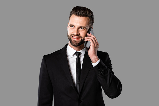 handsome smiling businessman in black suit talking on smartphone isolated on grey - Foto, immagini