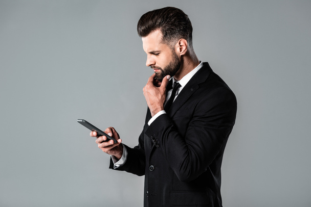 Seitenansicht des nachdenklichen Geschäftsmannes im schwarzen Anzug mit Smartphone isoliert auf grau - Foto, Bild
