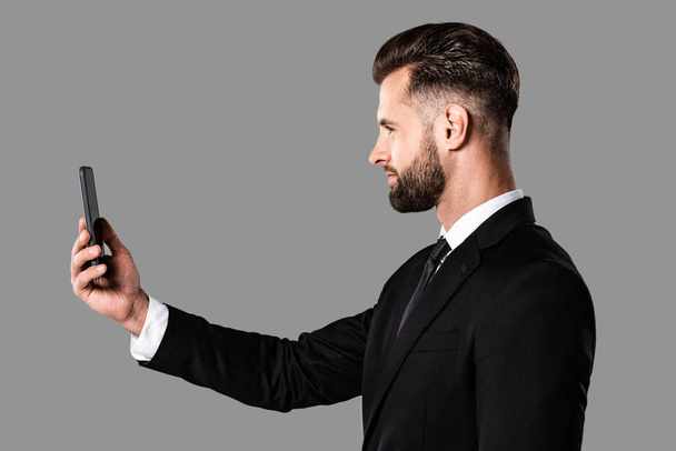灰色で隔離されたスマートフォンで自分撮りを撮る黒いスーツのハンサムなビジネスマンの側面図 - 写真・画像