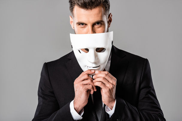 astuto hombre de negocios guapo en traje negro quitándose la máscara blanca aislado en gris
 - Foto, Imagen