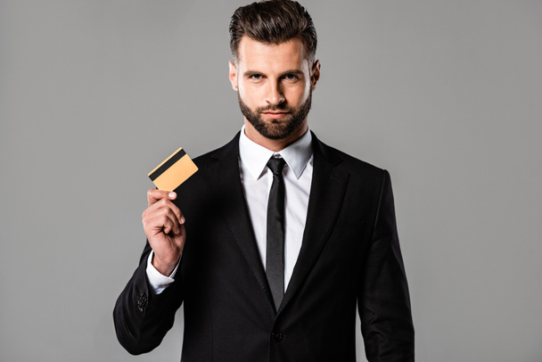 Красивый бородатый бизнесмен в черном костюме держит кредитку изолированной на сером
 - Фото, изображение