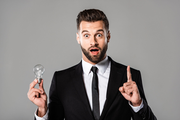shocked businessman in black suit holding light bulb and showing idea gesture isolated on grey - Foto, Imagen