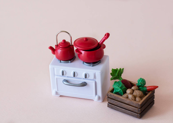 Mini-Figuren eines Teils der Küche: rote Teekanne und Topf mit Kochlöffel auf dem weißen Herd mit grauen Knöpfen und Knopf, neben Holzkiste mit Gemüse: Kartoffeln, Karotten, Kohl, Rüben - Foto, Bild