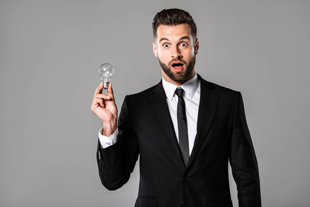 surprised businessman in black suit holding light bulb isolated on grey - Φωτογραφία, εικόνα
