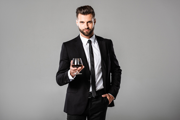 succesvolle zakenman in zwart pak houden van glas met whisky geïsoleerd op grijs - Foto, afbeelding