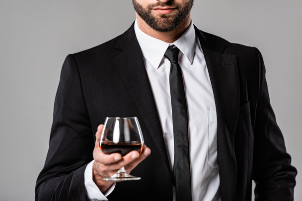 灰色で隔離されたウイスキーとガラスを保持する黒いスーツのひげを生やしたビジネスマンのトリミングされたビュー - 写真・画像