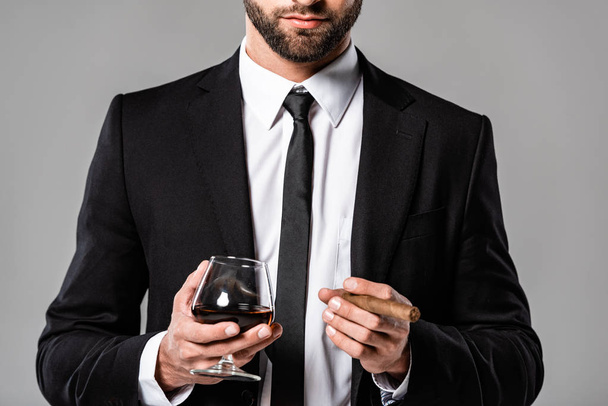 bijgesneden uitzicht van zakenman in zwart pak houden glas met whisky en sigaar geïsoleerd op grijs - Foto, afbeelding