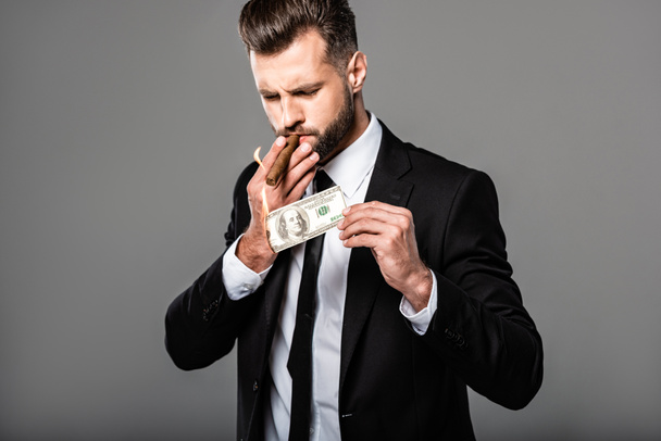 rich successful businessman in black suit lighting up cigar from burning dollar banknote isolated on grey - Photo, Image