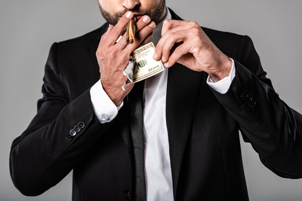 abgeschnittene Ansicht eines reichen Geschäftsmannes im schwarzen Anzug, der Zigarre von brennender Dollarnote isoliert auf grau anzündet - Foto, Bild
