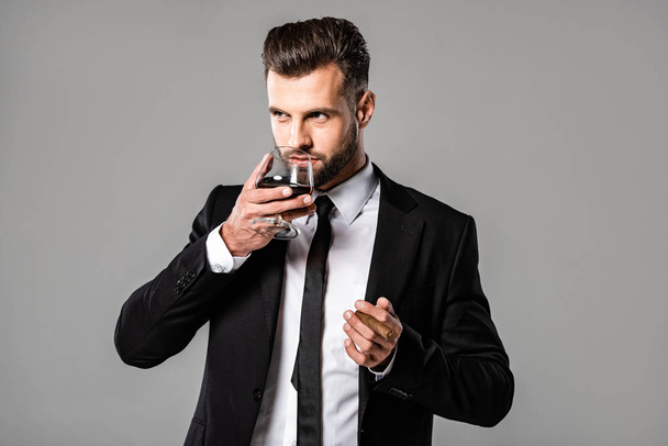 guapo hombre de negocios en traje negro bebiendo whisky aislado en gris
 - Foto, imagen