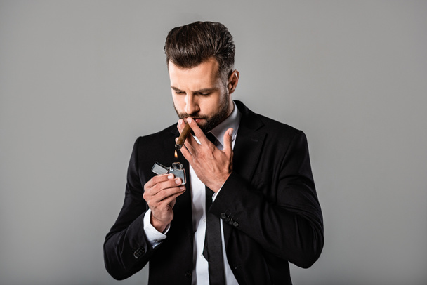 successful businessman in black suit lighting up cigar isolated on grey - Photo, Image