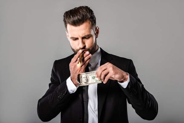 rijke succesvolle zakenman in zwart pak verlichting van sigaar van brandende dollar geïsoleerd op grijs - Foto, afbeelding