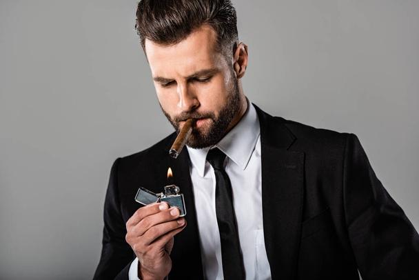 knappe zakenman in zwart pak verlichting van sigaar geïsoleerd op grijs - Foto, afbeelding