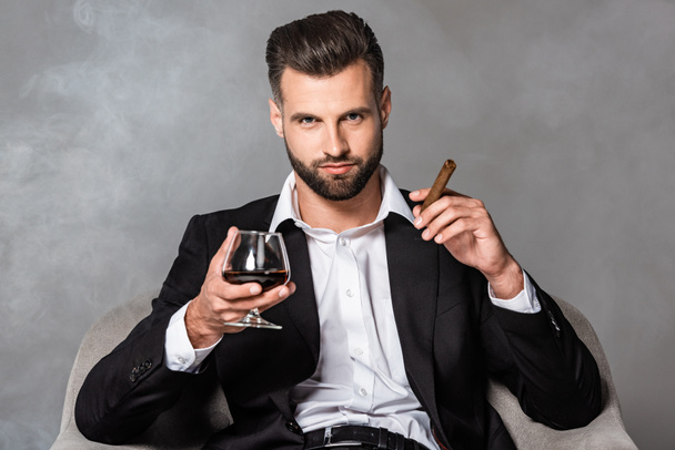 succesvolle zakenman in zwart pak zittend in fauteuil met sigaar en whisky in rook - Foto, afbeelding