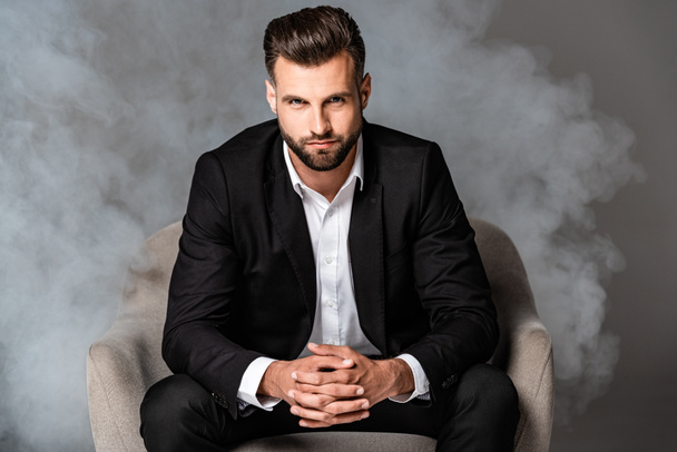 handsome businessman in black suit sitting in armchair in smoke on grey background - Photo, Image