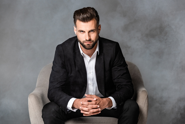 successful businessman in black suit sitting in armchair in smoke on grey background - Zdjęcie, obraz