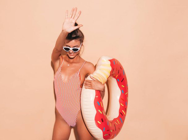 Joven hermosa mujer hipster sonriente sexy en gafas de sol. Chica en traje de baño de verano con colchón inflable donut lilo. Hembra positiva volviéndose loca. Cerca de la pared beige en gorra de visera transparente
 - Foto, Imagen