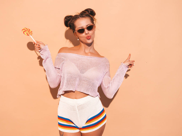 Closeup portrait of young beautiful sexy smiling woman with ghoul hairstyle. Trendy girl in casual summer white swimsuit in sunglasses.Hot model isolated on beige.Eating,biting candy lollipop - Fotó, kép