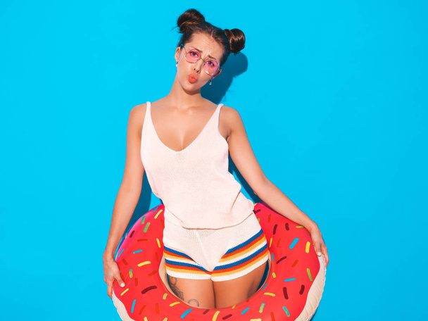 Young beautiful sexy smiling hipster woman in sunglasses.Girl in summer clothes with donut lilo inflatable mattress.Positive female going crazy.Funny model isolated on blue - Foto, Bild