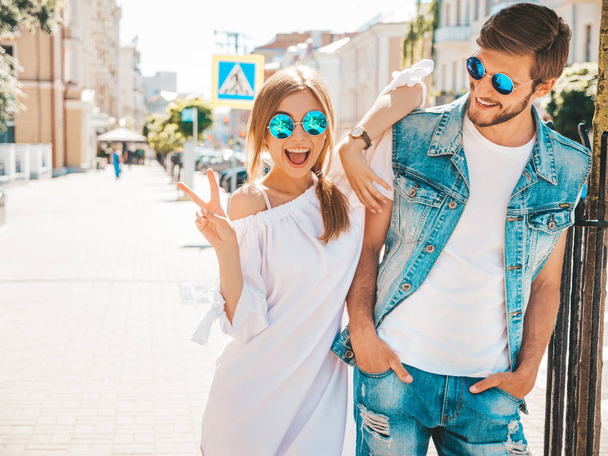 Smiling beautiful girl and her handsome boyfriend posing in the street. Woman in casual summer dress and man in jeans clothes. Happy cheerful couple family having fun in sunglasses - Foto, imagen