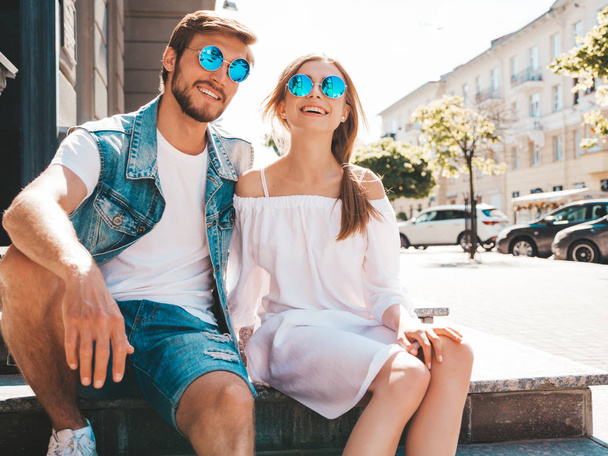 Smiling beautiful girl and her handsome boyfriend. Woman in casual summer dress and man in jeans clothes. Happy cheerful family. Sitting on stairs on the street background.Hugging couple - Foto, Bild